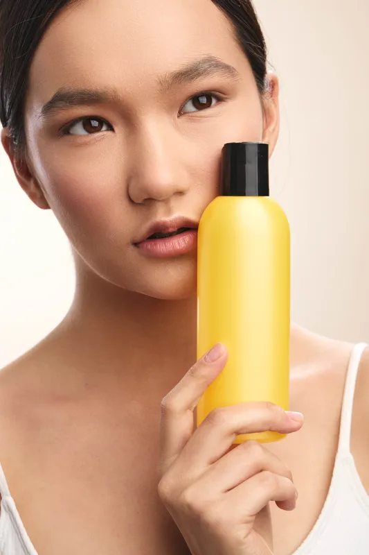 Asian woman holds a yellow bottle close to her face in a clean studio setting, presenting a skincare or beauty product with a calm, confident expression.