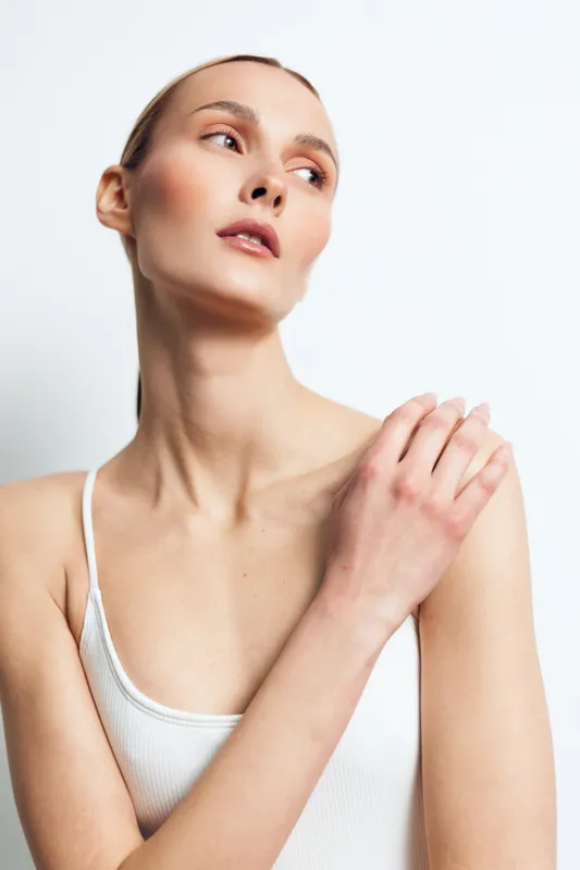 A young woman with clear skin and minimal makeup poses in a white tank top against a white background, highlighting natural beauty and skincare concepts.