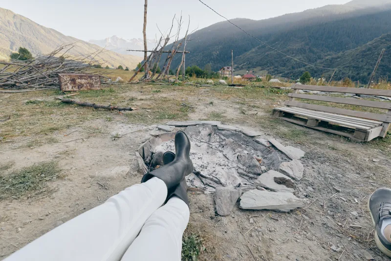 Boots legs campfire mountains bench ash outdoor countryside person relaxing at a cold fire pit on a rural campsite, feet up near wooden seating with valley view and worn grass under cloudy sky