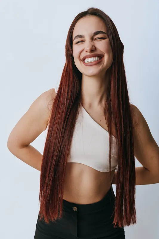 Young woman with long straight hair smiling with eyes closed wearing white crop top and black pants standing against plain background in casual style portrait.