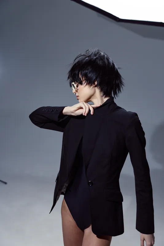 Stylish woman in black blazer and bodysuit posing with modern haircut and sunglasses in studio setting with gray background