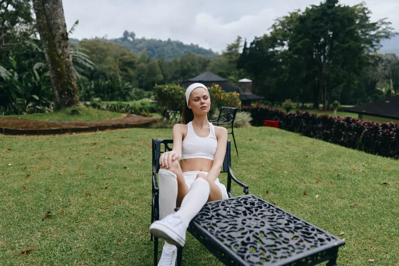 Young female model in sportswear relaxing outdoors amidst lush greenery and mountains, exuding a calm, confident vibe in an active lifestyle concept