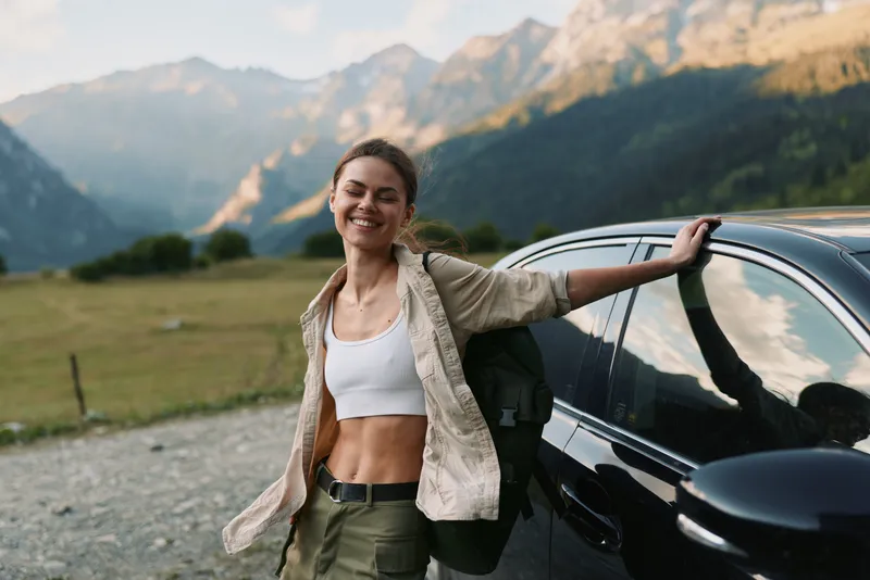 Woman enjoying outdoor lifestyle beside a car in mountain landscape, smiling confidently, wearing casual hiking attire, sunlit scenery and sense of adventure