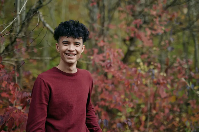 An authentic portrait of a young man outdoors, wearing a burgundy sweater, natural light, candid pose, and a believable, simple setting that conveys realism and trust.