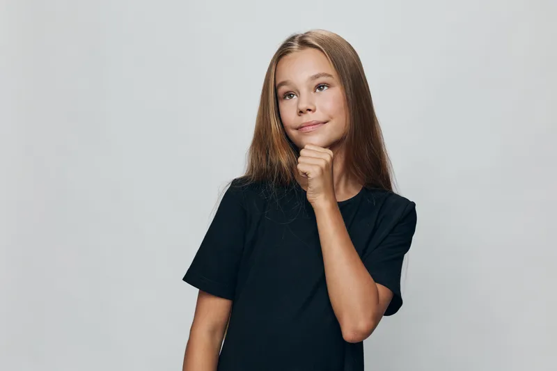 Girl with long hair in black t shirt posing thoughtful with hand near chin on gray background. Child standing and looking aside, casual portrait with natural expression.