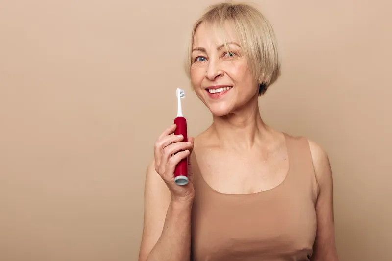 Smiling mature woman with short blonde hair holds electric toothbrush near face on beige background. Concept of dental care, oral hygiene, healthy smile, and personal grooming for seniors.