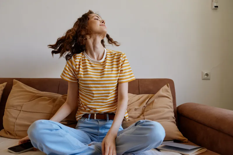 A woman lounging at home on a sofa, legs crossed, head tilted back with a carefree smile. Bright living room, striped tee and jeans. Concept for relaxation, selfcare and everyday comfort.