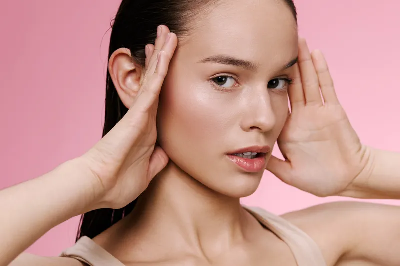 Beautiful young woman touching her face against a soft pink background, showcasing natural beauty, skin care and a healthy lifestyle