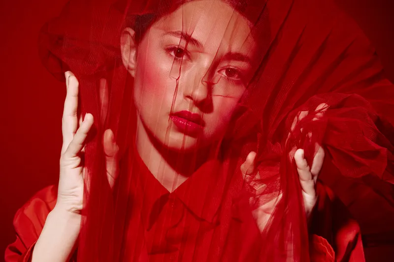 Woman in vibrant red costume poses skillfully behind red tulle fabric, creating a beautiful visual texture. Sharp eyes transform the composition into creative fashion and beauty art.