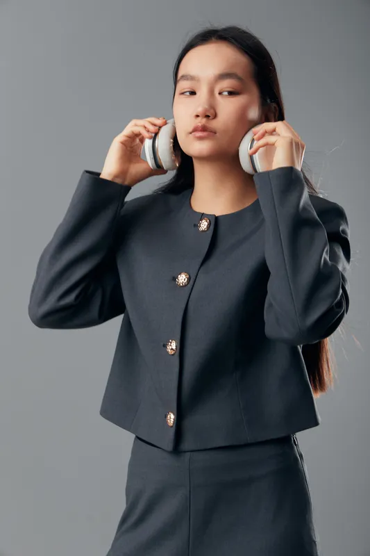 Young woman wearing formal jacket with buttons putting on wireless headphones standing over gray background. Asian female model with long hair and serious expression in studio portrait.