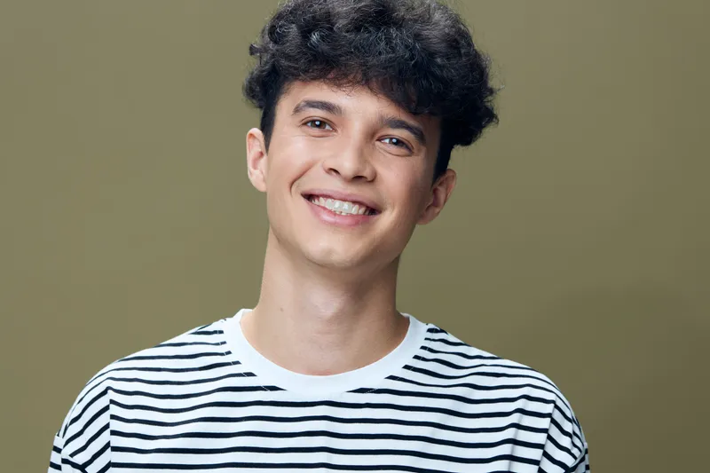 smiling man with curly hair wearing striped shirt, casual style, friendly expression, olive background, young adult, natural lighting portrait