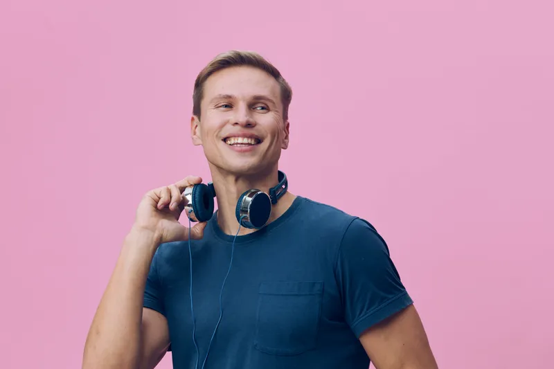 smiling young caucasian man wearing blue t-shirt holding headphones gesture looking aside over pink background digital detox concept