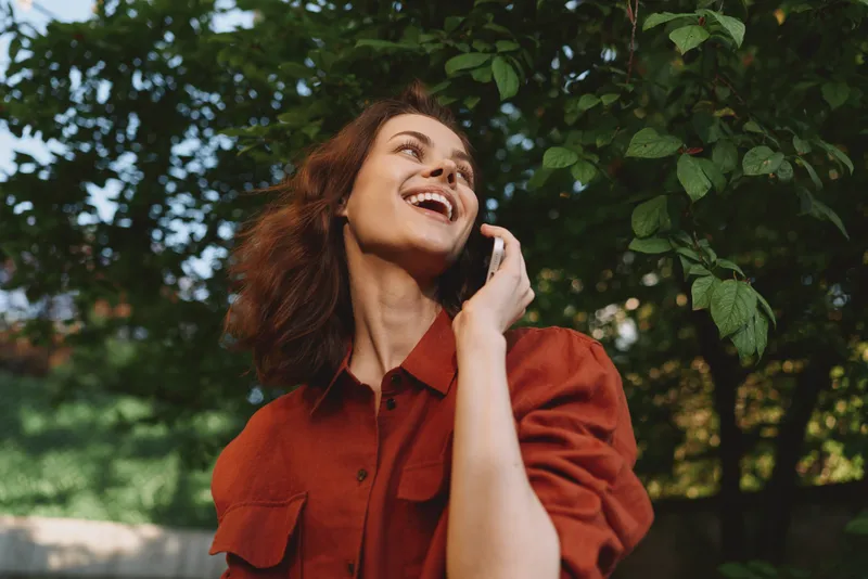 Smiling woman in a red shirt talking on a cellphone outdoors among green trees. Casual lifestyle moment with natural light, warm mood, and a candid, everyday feel