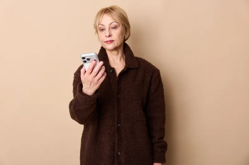 Smartphone, woman, blonde, portrait, communication, texting, messaging, technology, adult female holding mobile phone in dark brown winter coat indoors, beige background, calm attentive expression