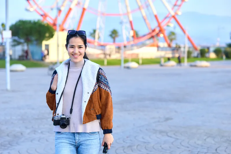 Traveler woman with camera in a sunny park near a towering ferris wheel, wearing a color block jacket and denim, capturing candid street moments and joyful energy under daylight.