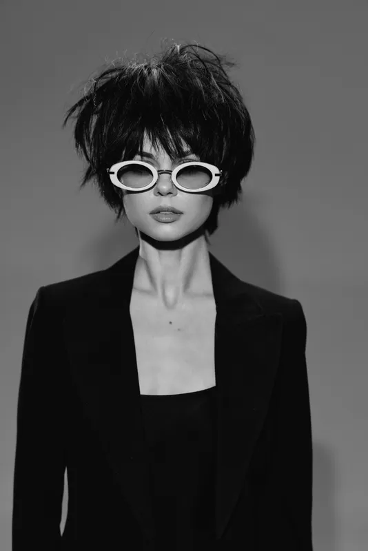 Black and white portrait of stylish woman with short hair wearing sunglasses and blazer, expressing cool and confident attitude.