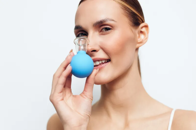 Young woman with blonde hair smiles gently while holding a blue glass bottle near her face against a white background in a clean skincare concept.