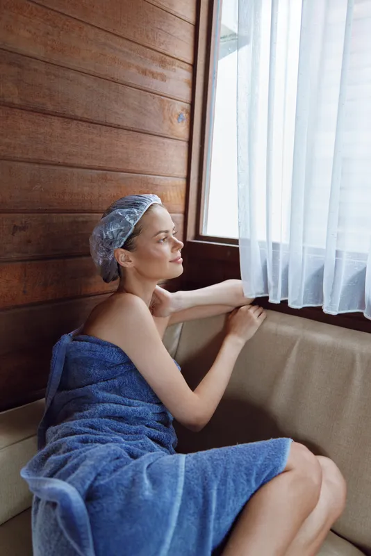 Relaxed woman wrapped in blue towel with shower cap sitting on wooden bench, enjoying natural light through white curtains in cozy sauna interior.