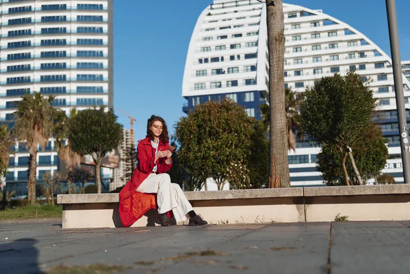 A young woman in a red coat sits on a concrete bench outdoors using a smartphone on a sunny day with modern buildings and trees in the background.