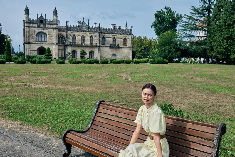 Woman in a pale dress sits on a wooden bench in a sunlit park, with a historic castle silhouette in the background, creating a serene and elegant outdoor scene.