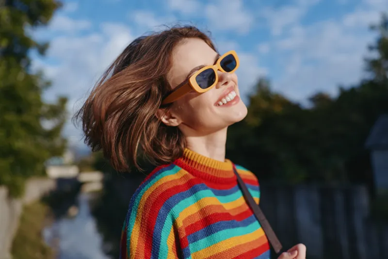 Young woman in her mid twenties wearing a rainbow sweater and yellow sunglasses, smiling outdoors by a canal with casual bag, windblown hair and sunny travel lifestyle joyful mood.
