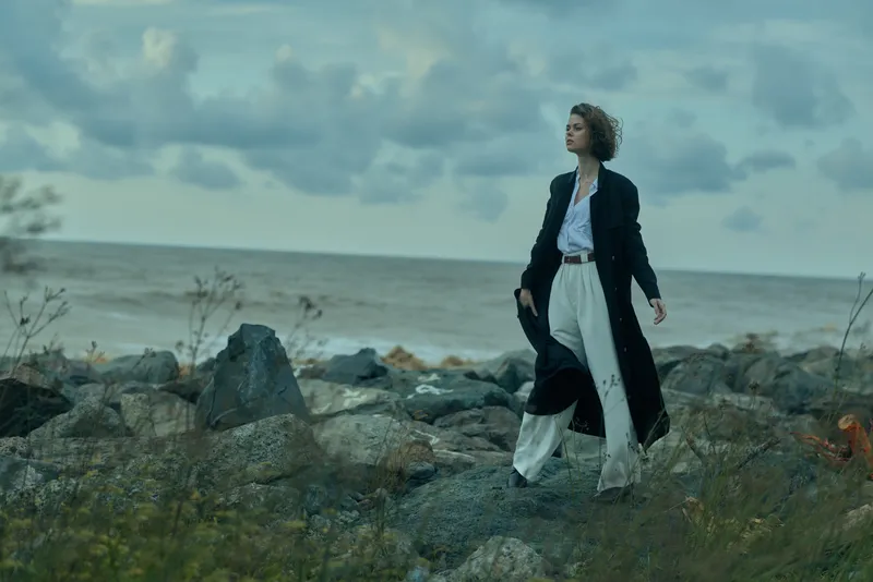 Coast scene with a woman walking along rocky shoreline, wind sweeping her long coat and flowing dress, seascape backdrop, moody clouds, dramatic atmosphere, travel mood, minimal fashion against nature