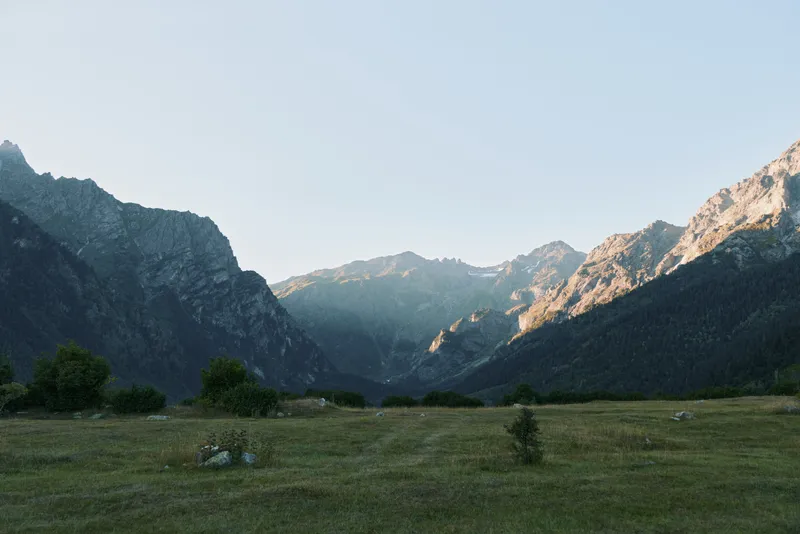 A broad mountain landscape unfolds over a grassy valley under a clear sky. Rugged slopes frame the scene, inviting reflection and exploration in a peaceful outdoor setting.