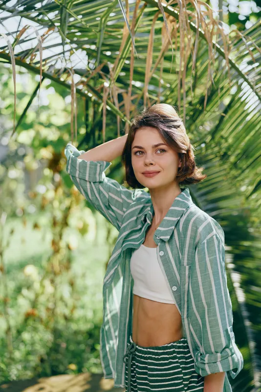 Portrait of a young woman outdoors in a striped green shirt and white crop top amid lush tropical greenery.