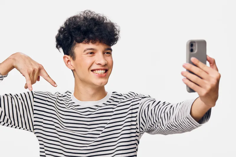 smiling man taking selfie with smartphone pointing finger, casual striped shirt, curly hair, isolated white background, cheerful and youthful expression