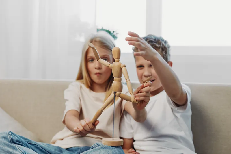 Two children sitting on a couch play with a wooden artist mannequin, focusing on creativity and childhood imagination in a bright, casual home setting.