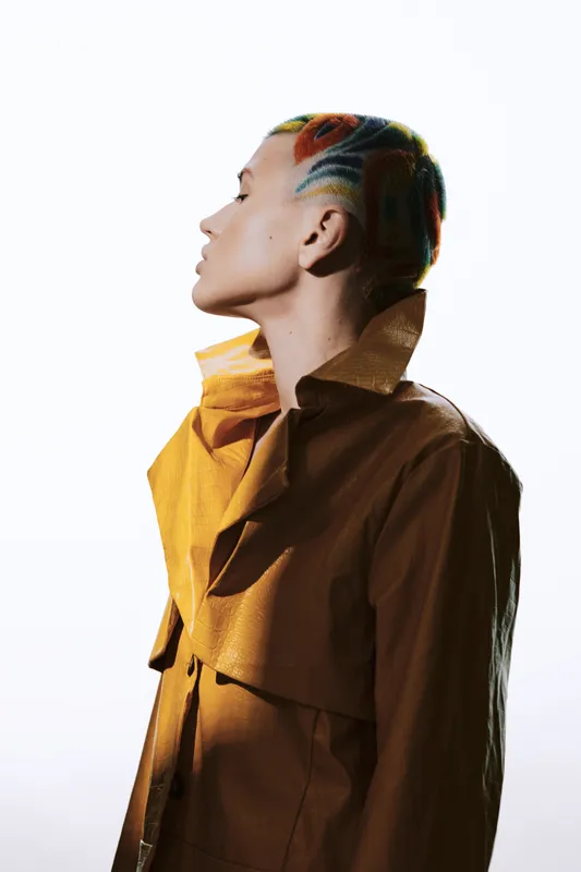 Woman with colorful braided hair wearing a stylish brown coat in a studio setting, showcasing creative fashion and unique hairstyle art.