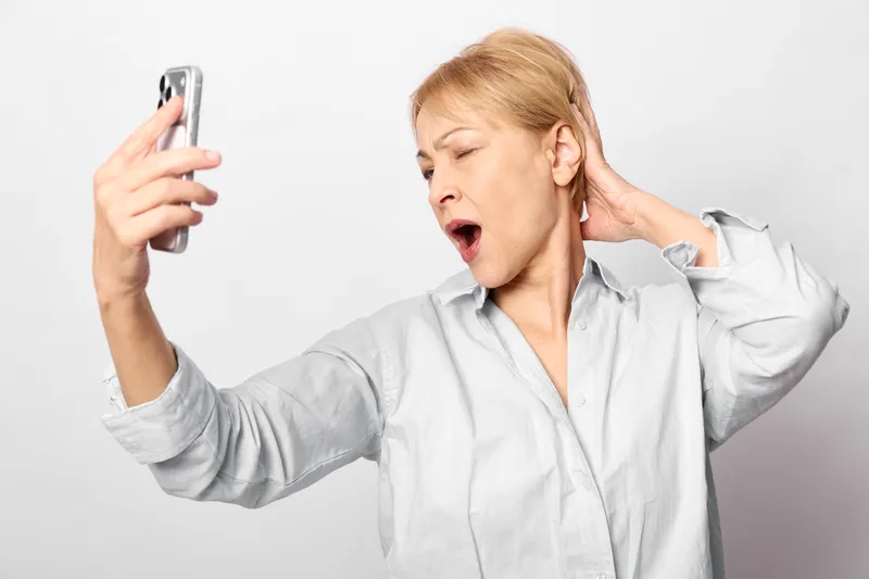 Woman smartphone selfie pose, shouting with stressed face, hands on ears, communication noise frustration overload, concept mental health, white studio