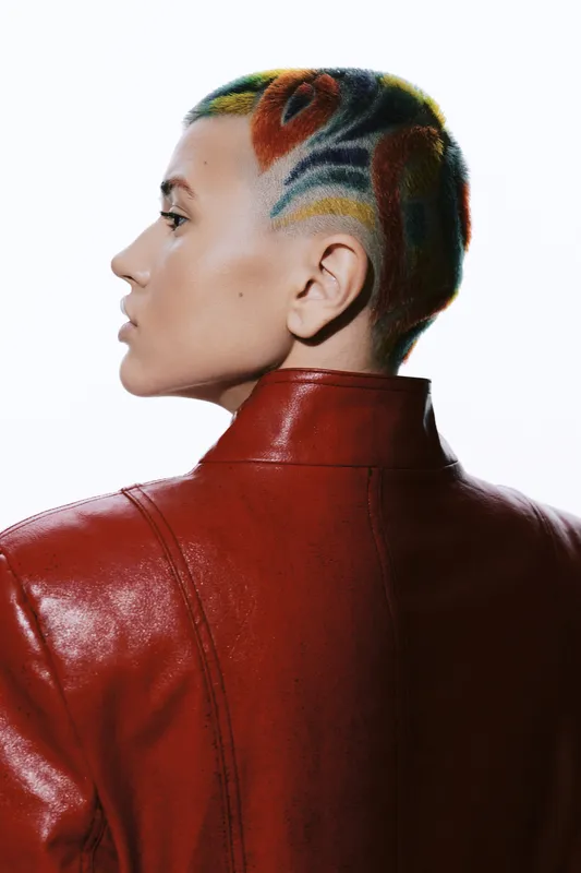 Creative woman with colorful shaved hair wearing a red leather jacket, showcasing unique style and confident attitude in a modern studio setting.