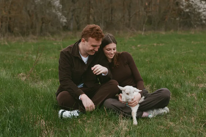 Casual couple sits on grass in a park with a small goat kid, sharing a warm smile, relaxed mood, and gentle touch, natural backdrop, rustic charm, affectionate moment, lifestyle vibe, peaceful