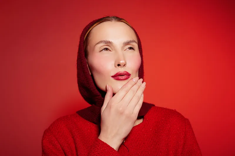 Young woman in red sweater and headscarf poses thoughtfully on red background. Concept of fashion, style, and winter clothing with bright makeup and confident expression.