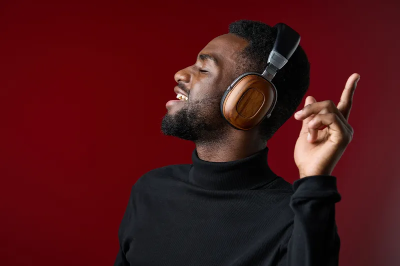 Young Black man enjoying music with wooden headphones, smiling joyfully against a vibrant red background, exuding happiness and style
