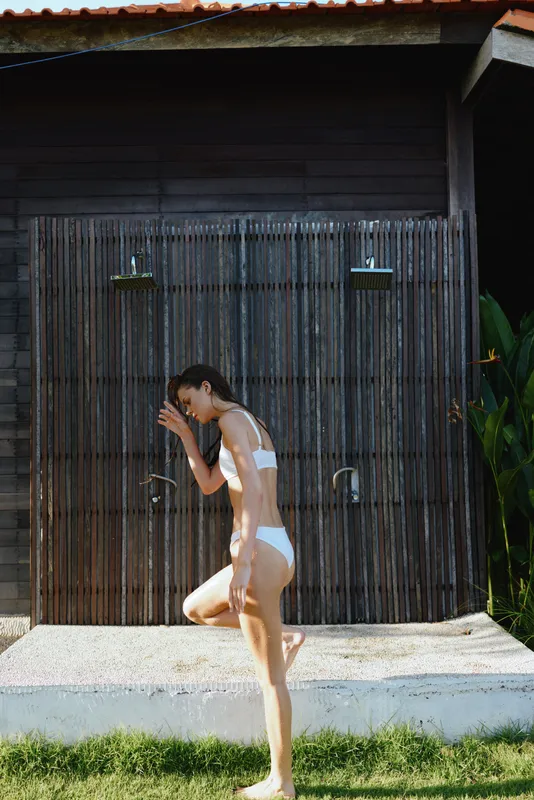 A woman in a white swimsuit runs under outdoor showers beside a wooden shed, a playful summer moment captured in bright light. A lifestyle scene focused on outdoor activity, water, sun, and carefree