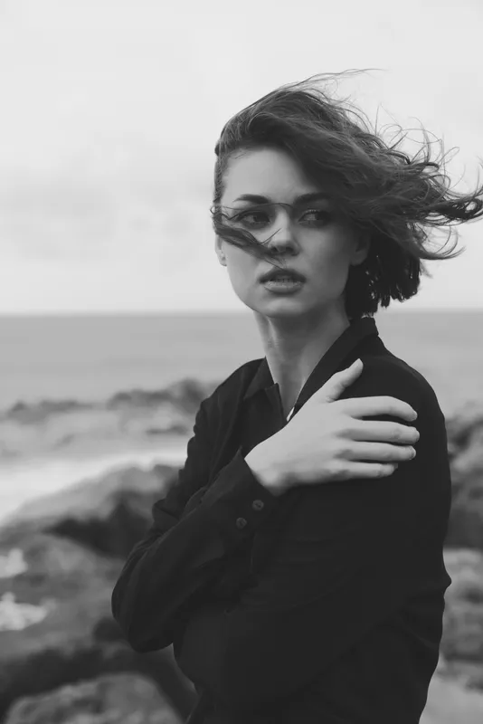 Moody monochrome photo of a woman by the sea, windblown hair and contemplative gaze, coastal fashion mood with dramatic lighting and ocean breeze