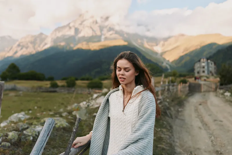 A young woman with brown hair walks along a rustic fence beside a mountain tapestry wrapped in a light knit sweater calm and thoughtful in the open countryside