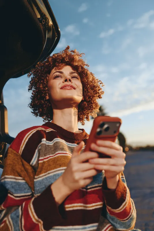 Young woman lifestyle inside car, curly hair woman in striped sweater, holding smartphone and looking up, warm sunset light, outdoors road and sky background, casual travel