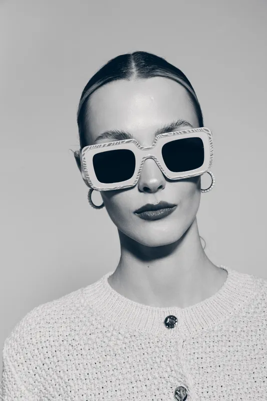 Stylish white woman with pale skin wearing large sunglasses and hoop earrings in a white textured sweater. Modern fashion portrait with minimalist background and confident expression.
