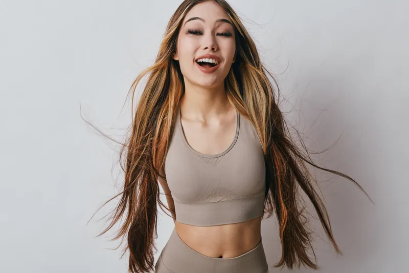 Young woman with long hair in beige sportswear posing and smiling with eyes closed. Fitness, wellness and healthy lifestyle with happiness and confidence in studio.