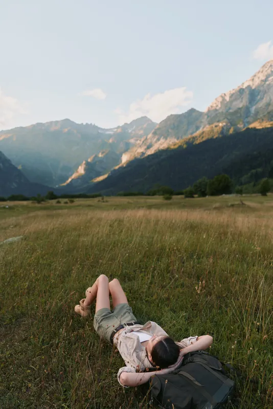 A woman lies on her back in a green meadow surrounded by mountains under a clear sky, enjoying a peaceful moment in nature during a sunny day.