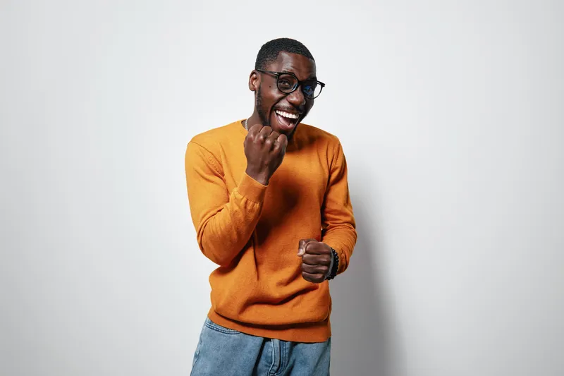African American man with dark skin wearing glasses and orange sweater smiling and celebrating success with clenched fists on white background. Casual style and positive emotion.