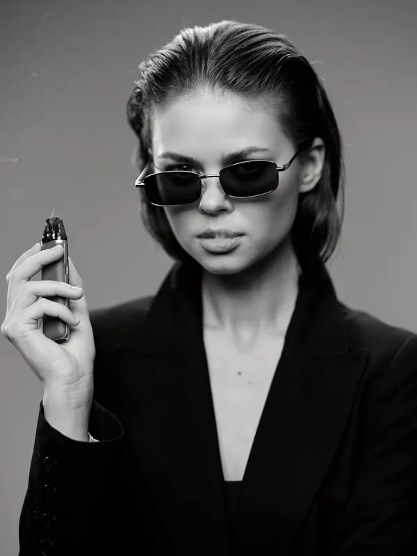 Stylish woman wearing sunglasses and black blazer holding vape device in black and white portrait