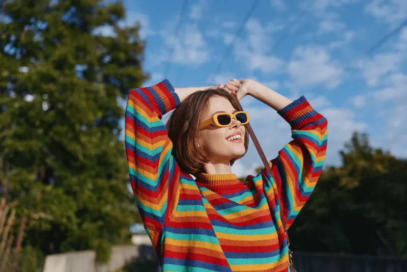 Young woman in a rainbow sweater and yellow sunglasses smiling in an outdoor portrait, joyful gesture with arms raised, looking up in sunny park for lifestyle fashion.