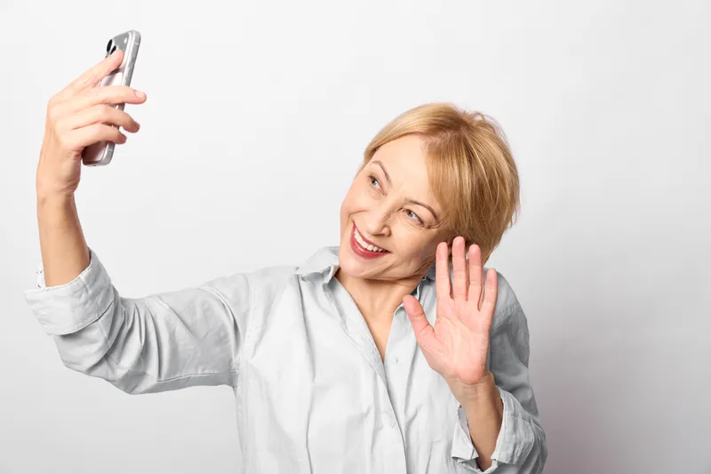 Smiling woman holding a smartphone for a selfie, waving hand beside her face in a bright studio, white portrait background, communication and technology concept