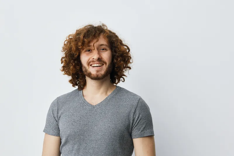 Cheerful young man with curly hair smiling on a neutral background, showcasing casual style and warm emotions, perfect for lifestyle or personal branding use