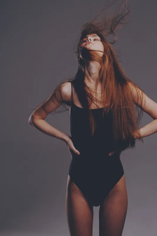 Woman with long hair wearing black bodysuit posing with eyes closed and hair blowing, expressing freedom and confidence in studio lighting.
