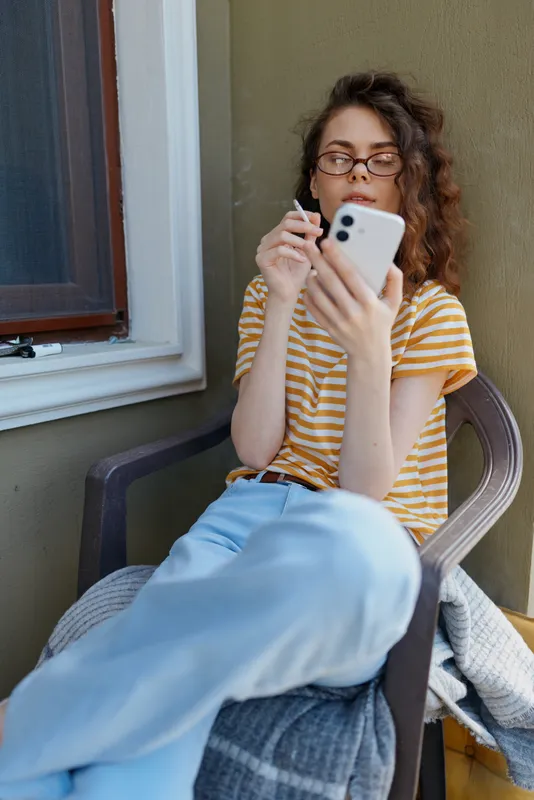 A relaxed woman sits on a porch chair, scrolling on a smartphone, wearing a striped tee and jeans. Bright natural daylight frames a casual home setting, highlighting everyday connectivity and calm.